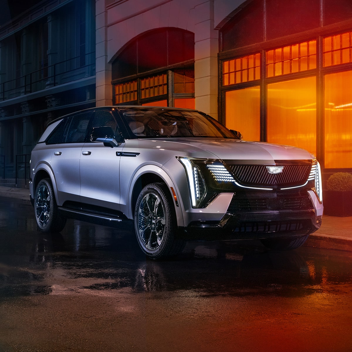 Front-side view of the 2025 Cadillac Escalade IQ electric suv parked on wet flooring outside a brightly-lit building.