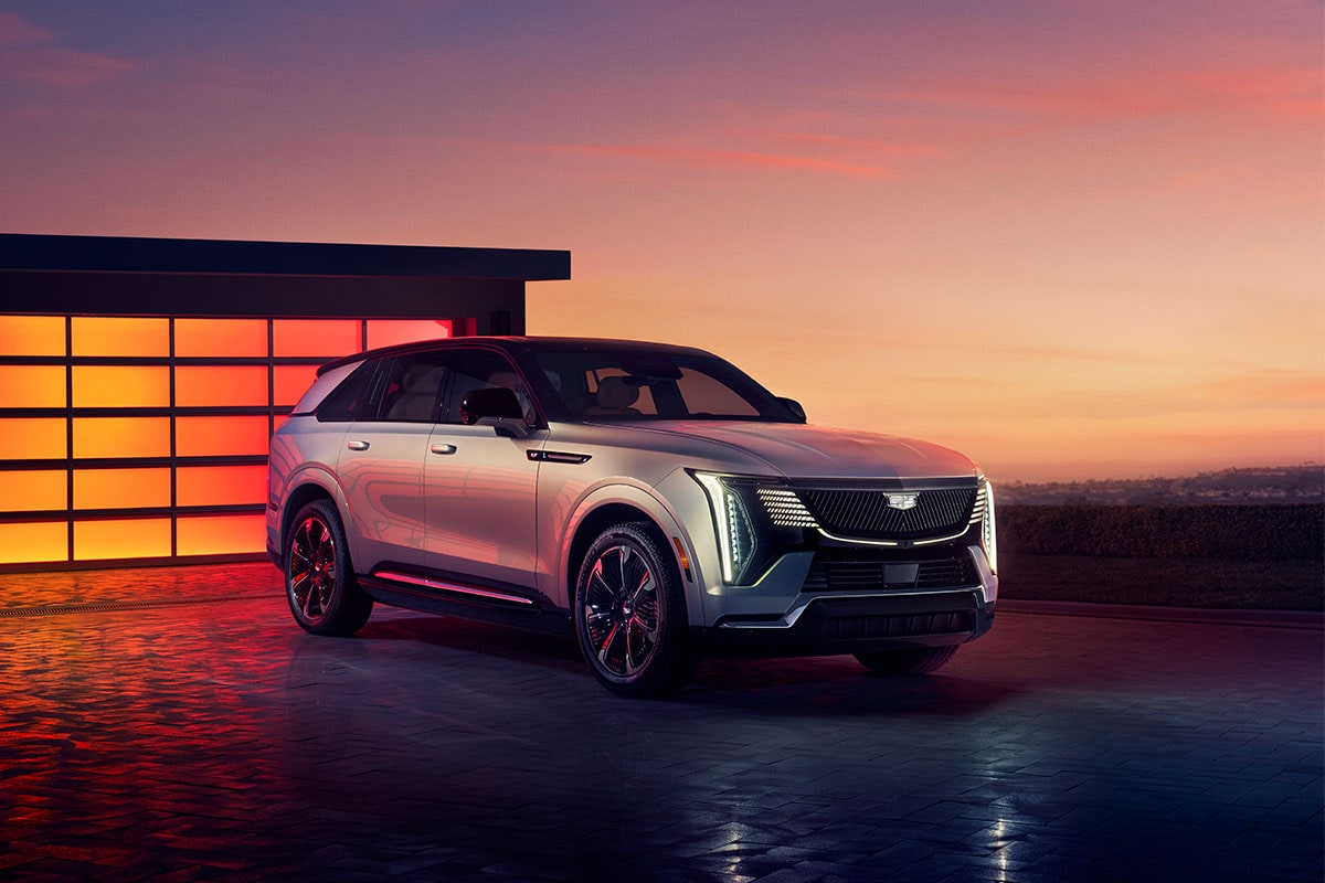 Front-side view of the 2025 Cadillac Escalade IQ electric suv parked outside a brightly-lit modern home during sunset.