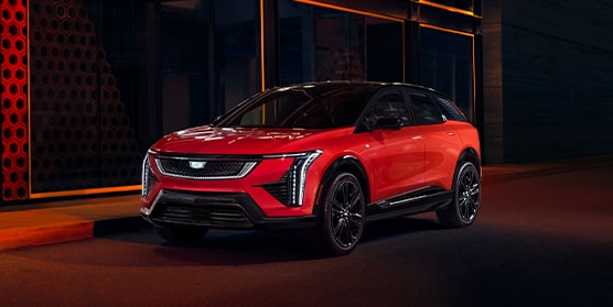 Front-side view of the 2025 Cadillac OPTIQ parked beside a brightly-lit sidewalk at night.